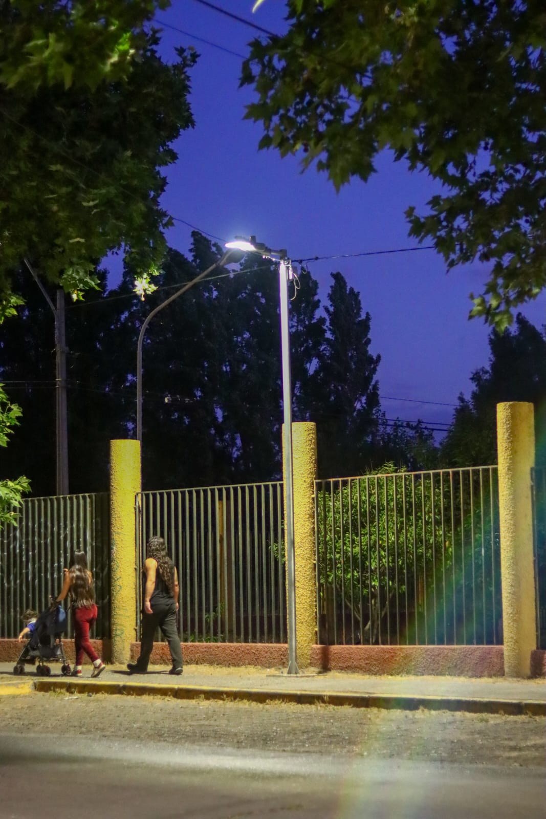 Luminaria peatonal encendida en calle San Alfonso, mientras dos vecinas caminan por la vereda iluminada.